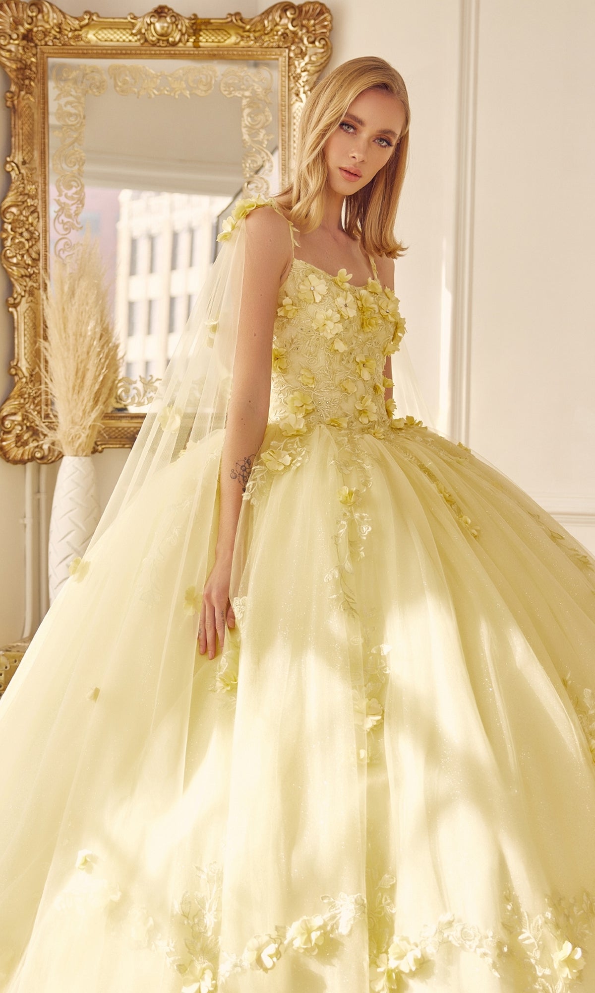 A woman in the Juliet 1436 Glitter Quinceañera Dress with Cape stands indoors before an ornate gold-framed mirror.